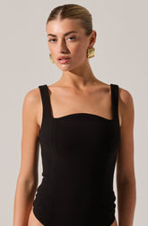 A woman is wearing a black sleeveless top with wide straps that sit on her shoulders. The top has a square neckline with a slight curve across the bust. She is accessorized with large, gold, spiral-shaped earrings.
